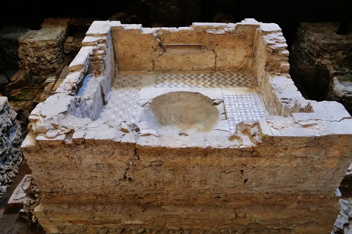 A square stone basin with a rounded hollow at its centre, edged with pale tesserae, set in dark excavated ground beneath a church