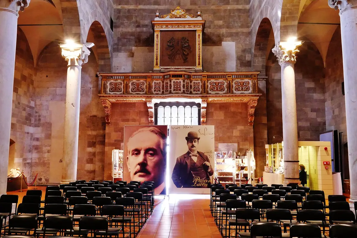 Rows of black chairs filling the floor of a Romanesque nave in Lucca, with a gilded organ loft at the far end and two large portraits of Puccini on boards below it