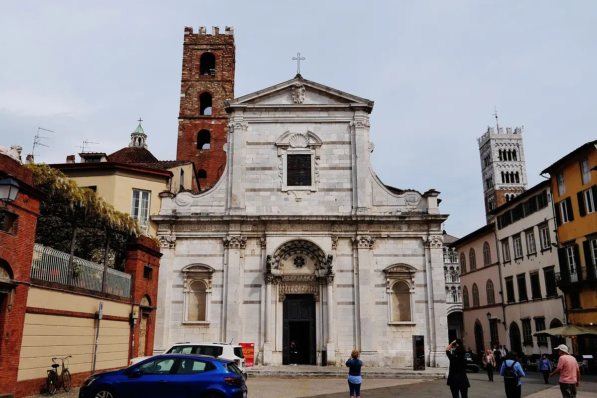 The pale marble front of a Lucca church with a brick bell tower behind it and the striped cathedral tower on the right.