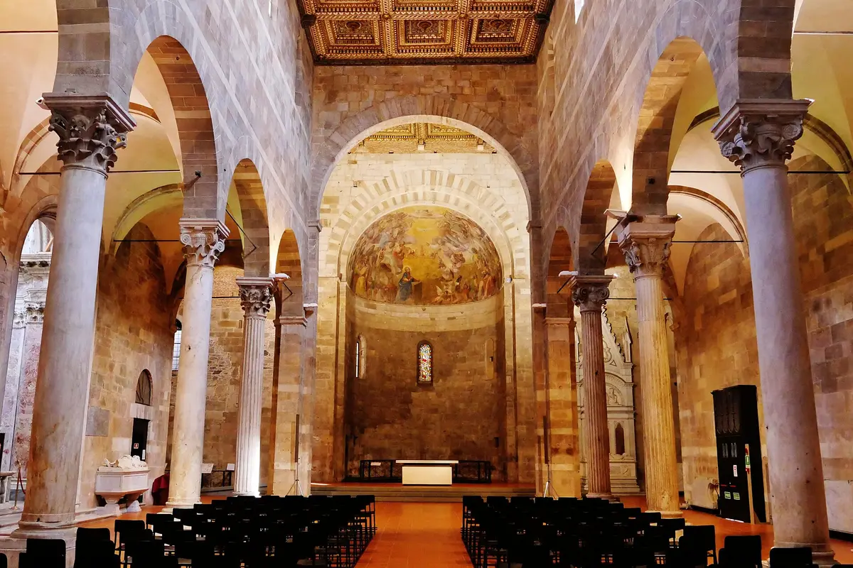 A long Romanesque nave with rows of black chairs facing a painted apse at the far end, between stone arches and tall columns.