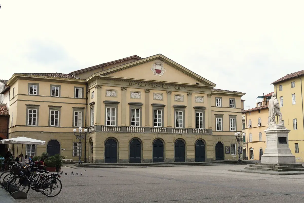 The pale yellow neoclassical front of the Teatro del Giglio in Lucca, with a row of arched doorways at street level and a triangular pediment above, seen across an open square